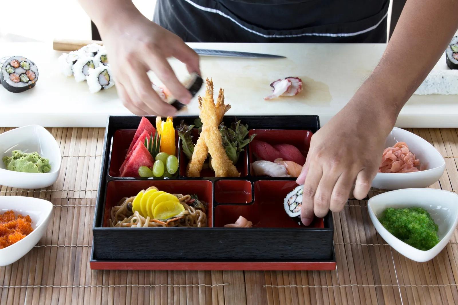 A chef assembling a sushi and sashimi bento box, condiments arranged in small dishes around it