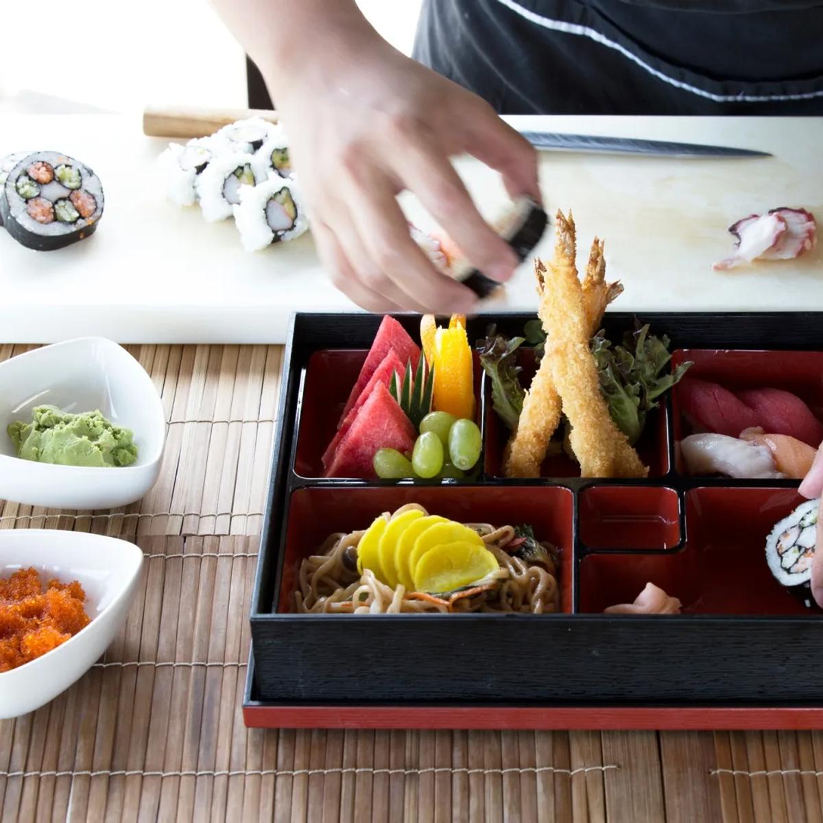A chef assembling a sushi and sashimi bento box, condiments arranged in small dishes around it