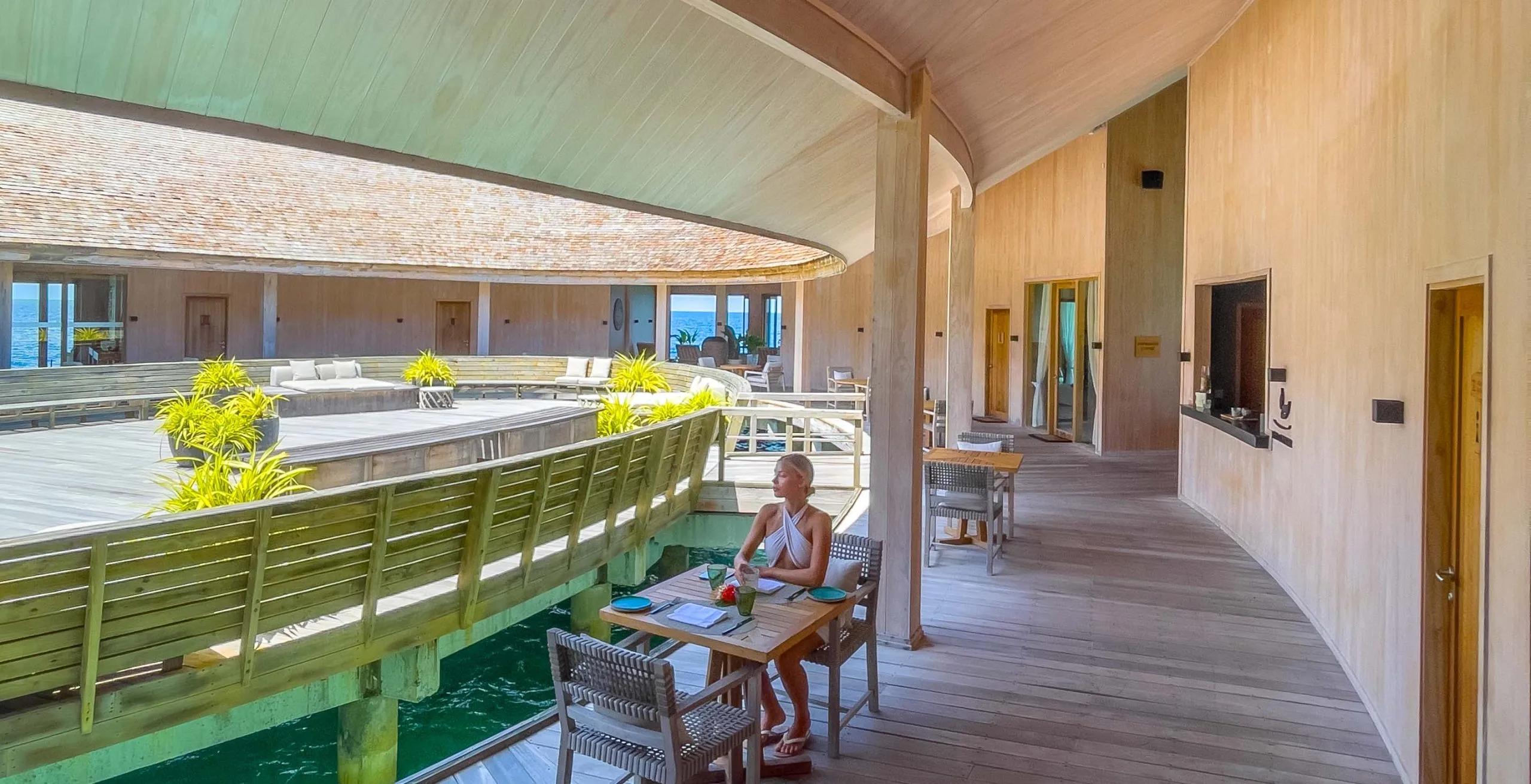 A guest seated at a table on an overwater walkway between guest room wings, green lagoon water visible below
