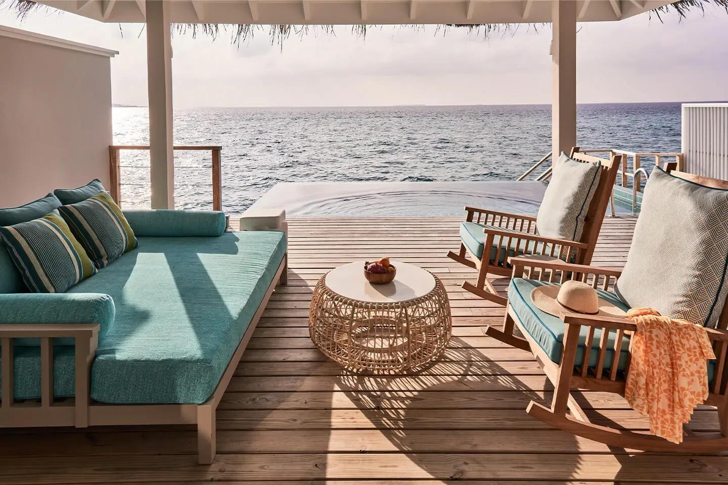 An Ocean Pool Villa's overwater-style deck, its tiled infinity pool appearing to merge with the sea beyond, two rocking chairs set under the thatched roof
