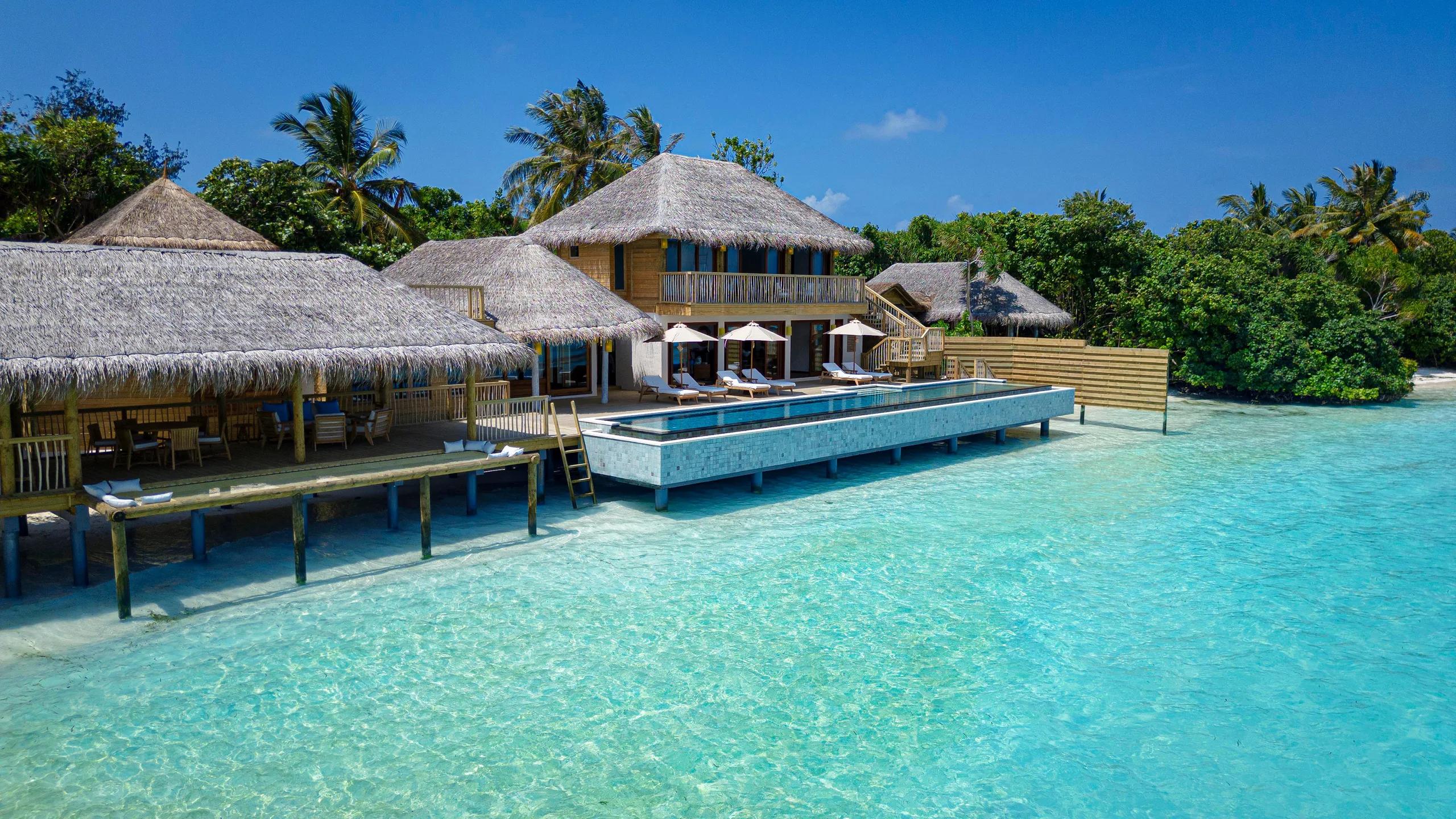 A large beachfront villa complex with multiple thatched roofs, sun loungers and umbrellas along a long pool deck