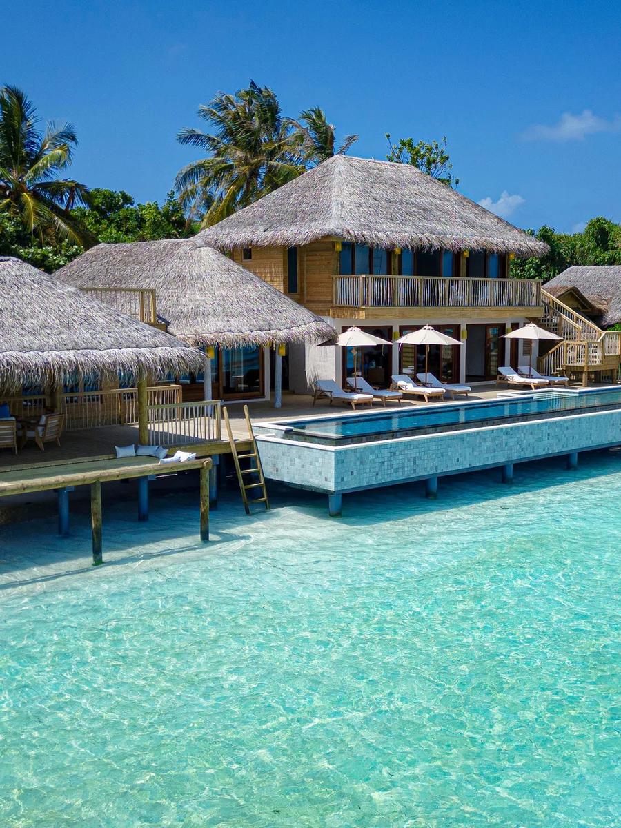 A large beachfront villa complex with multiple thatched roofs, sun loungers and umbrellas along a long pool deck