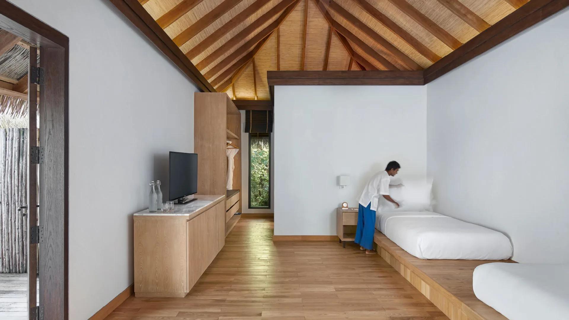 Staff member arranging pillows on a twin bed in a wood-panelled bedroom with a thatched ceiling