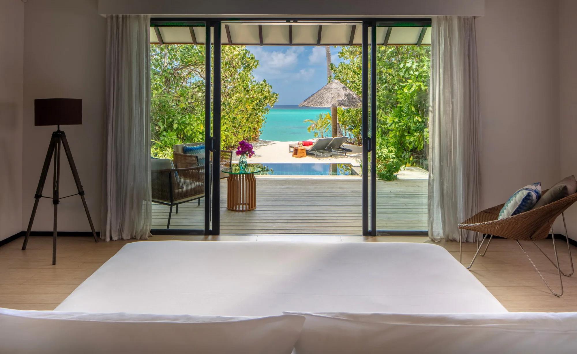 A Sunrise Beach Pool Villa's deck seen through open sliding doors, with a thatched umbrella, sun loungers and a private pool leading toward the lagoon