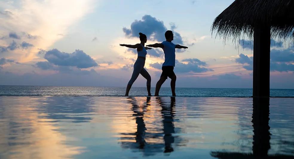 Two silhouetted figures in a lunging pose at the edge of an infinity pool, sunset colours reflected in the still water