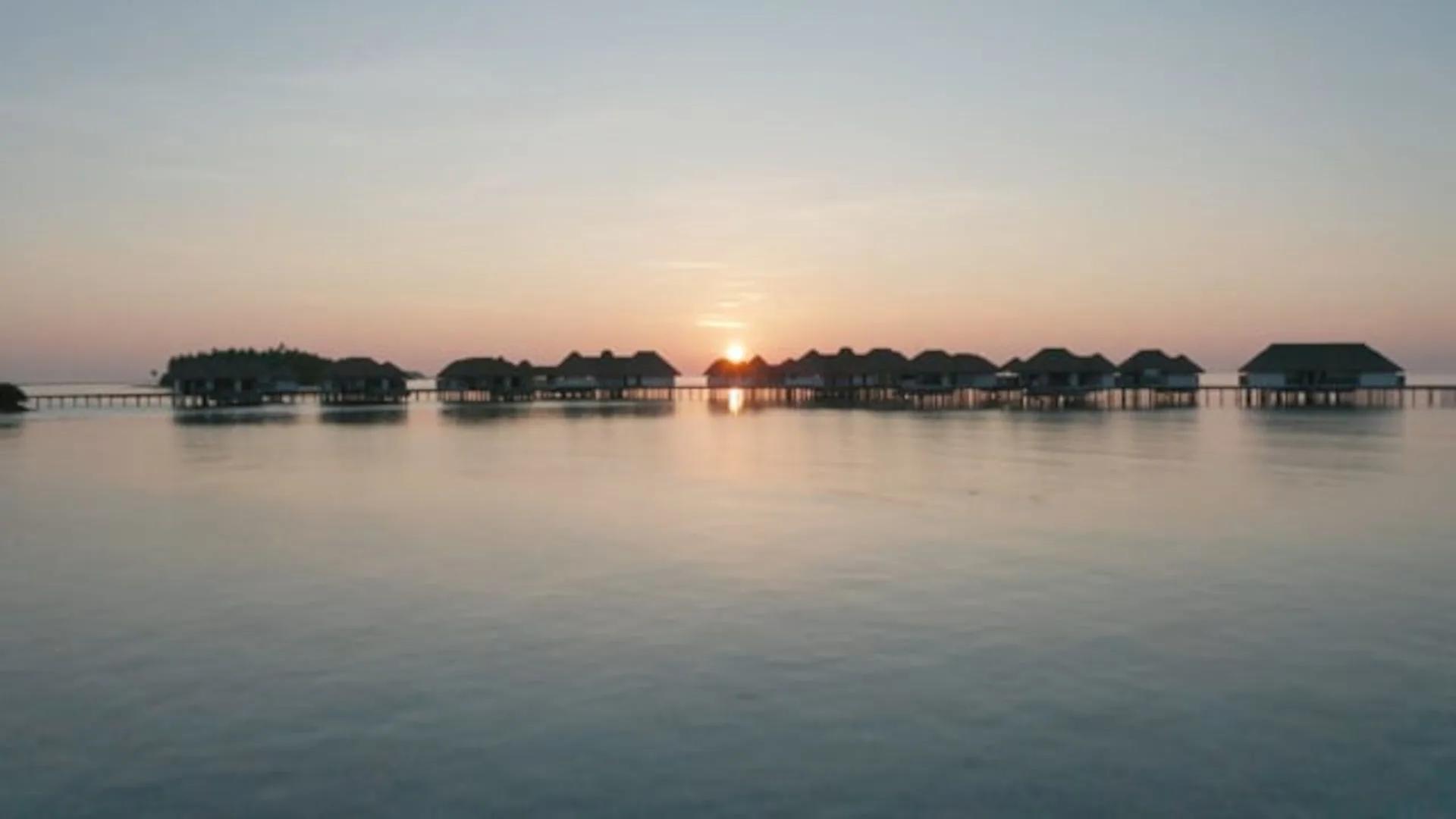 Overwater villas silhouetted against a pink sunset sky, seen across the still lagoon