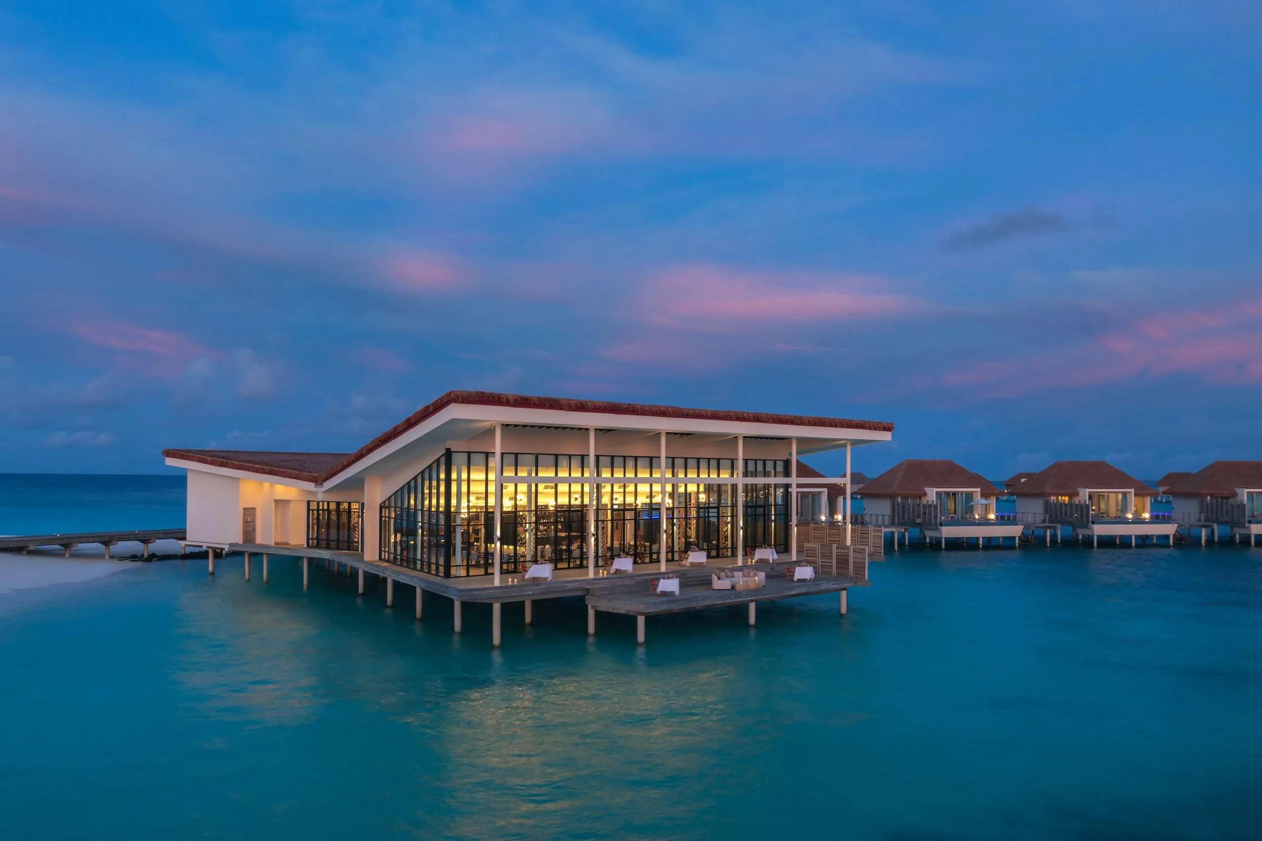 Kabuki's overwater dining pavilion at Radisson Blu Resort Maldives lit up at dusk, other overwater villas visible along the jetty behind it