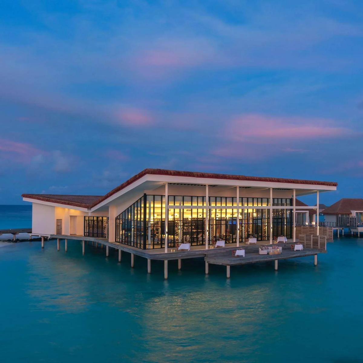 Kabuki's overwater dining pavilion at Radisson Blu Resort Maldives lit up at dusk, other overwater villas visible along the jetty behind it