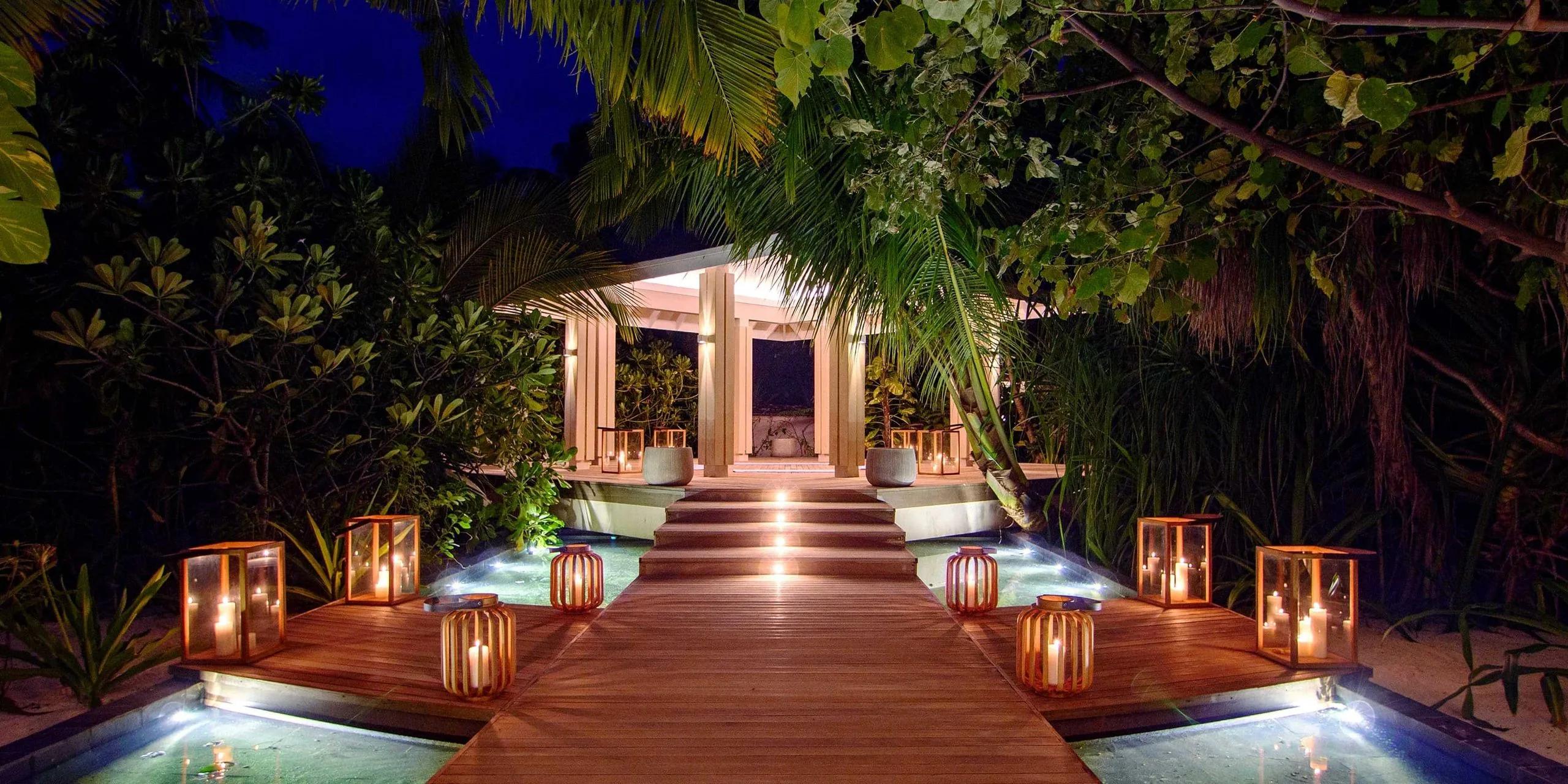 A lantern-lit timber walkway across a reflecting pool leading to the Aura Spa pavilion at Baglioni Maldives after dark