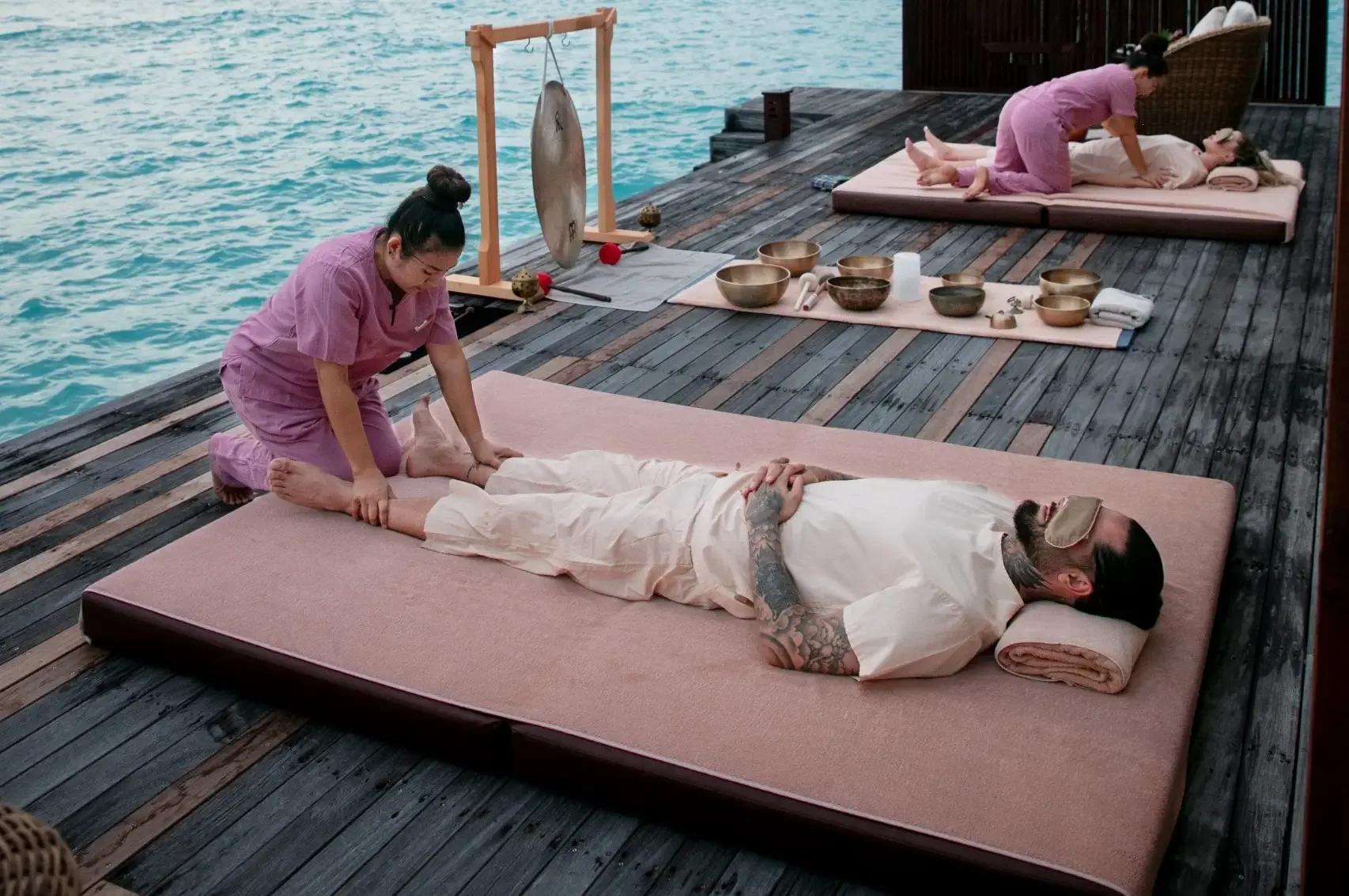 Therapists giving an overwater massage treatment at The Spa Retreat, singing bowls set out on the deck beside the lagoon