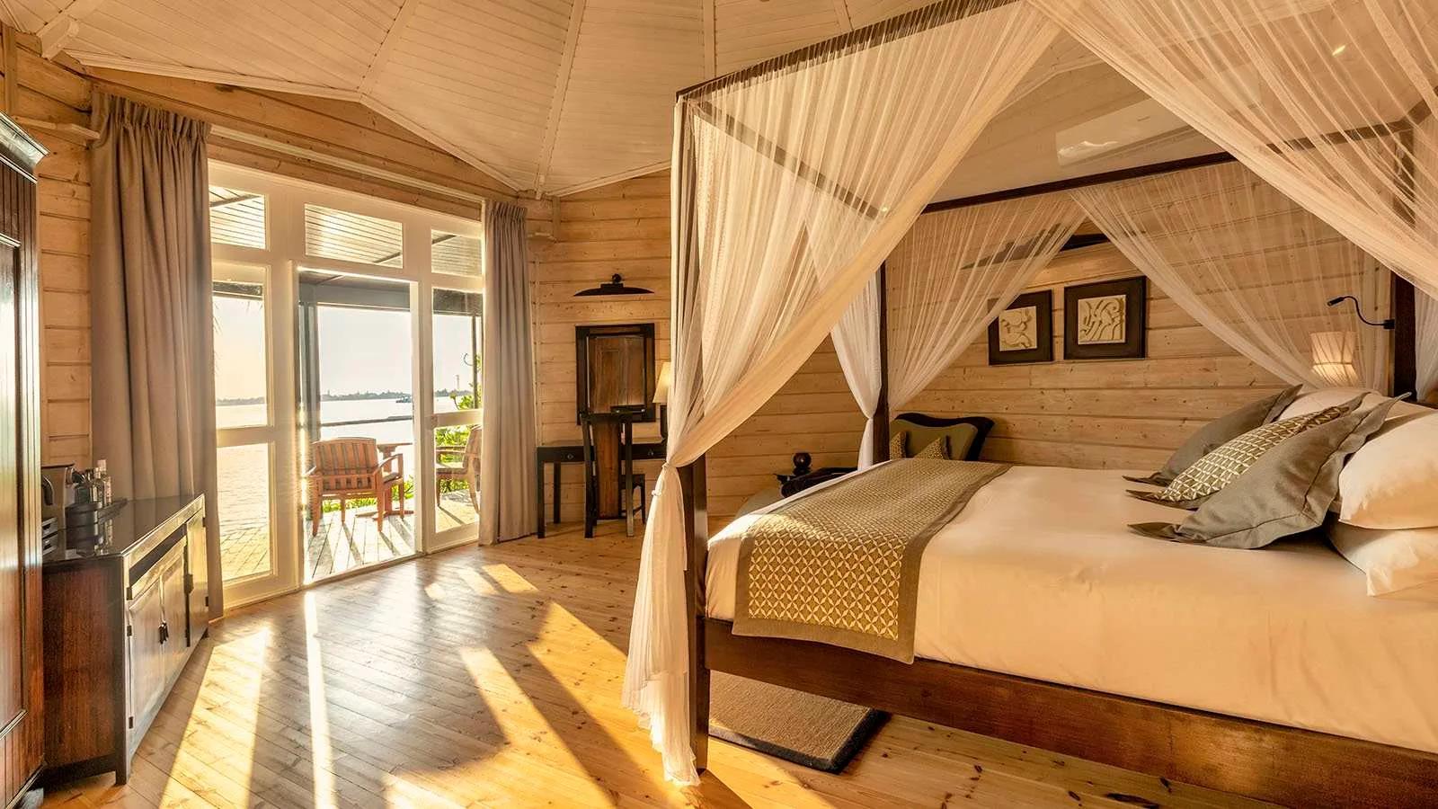 The interior of a Premium Jacuzzi Beach Villa, a four-poster bed beneath a peaked timber ceiling with the beach visible through the open doors