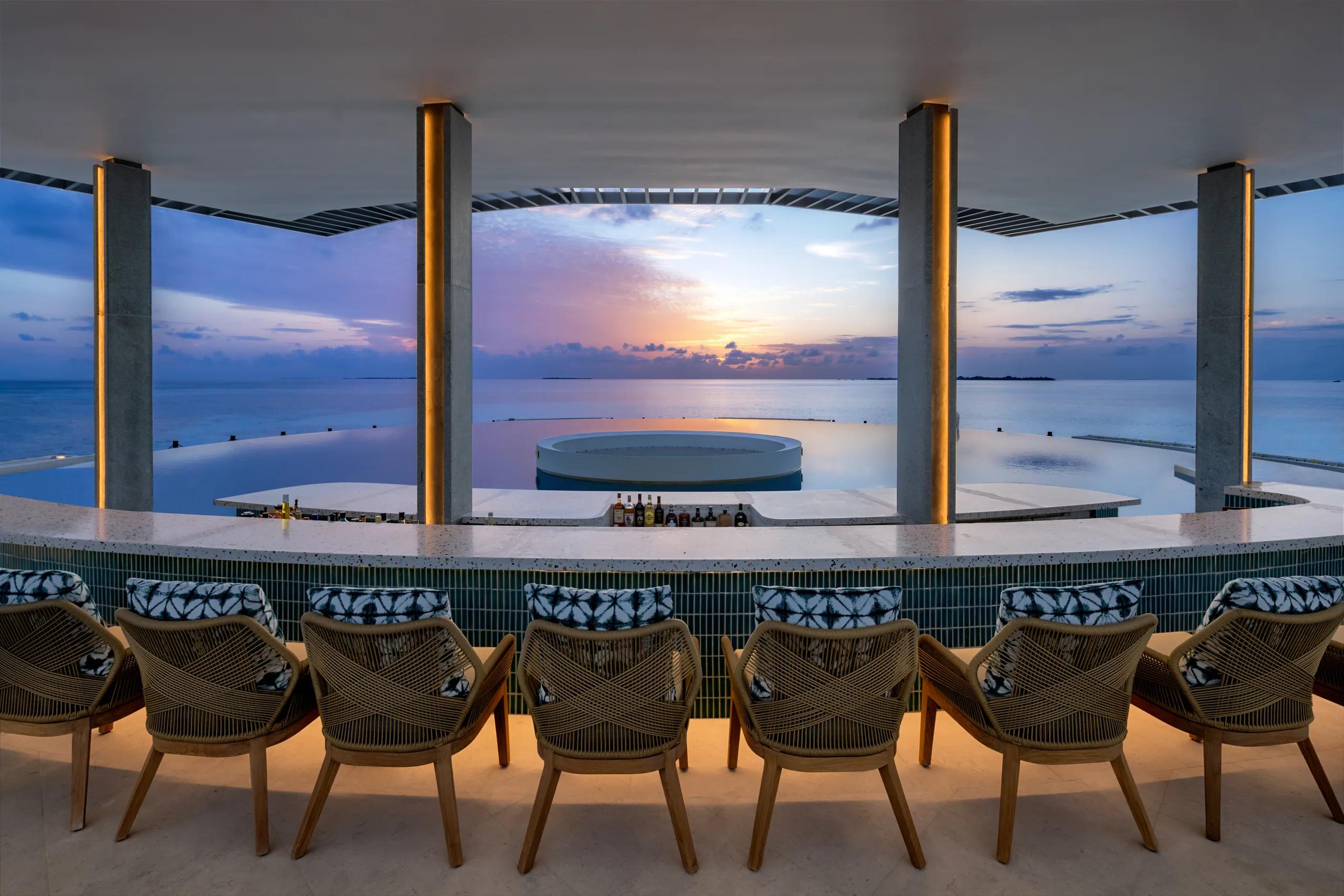 A rooftop bar at sunset, with a curved bar counter, woven armchairs, and an infinity pool overlooking the ocean.