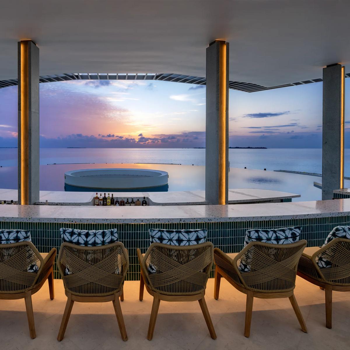 A rooftop bar at sunset, with a curved bar counter, woven armchairs, and an infinity pool overlooking the ocean.