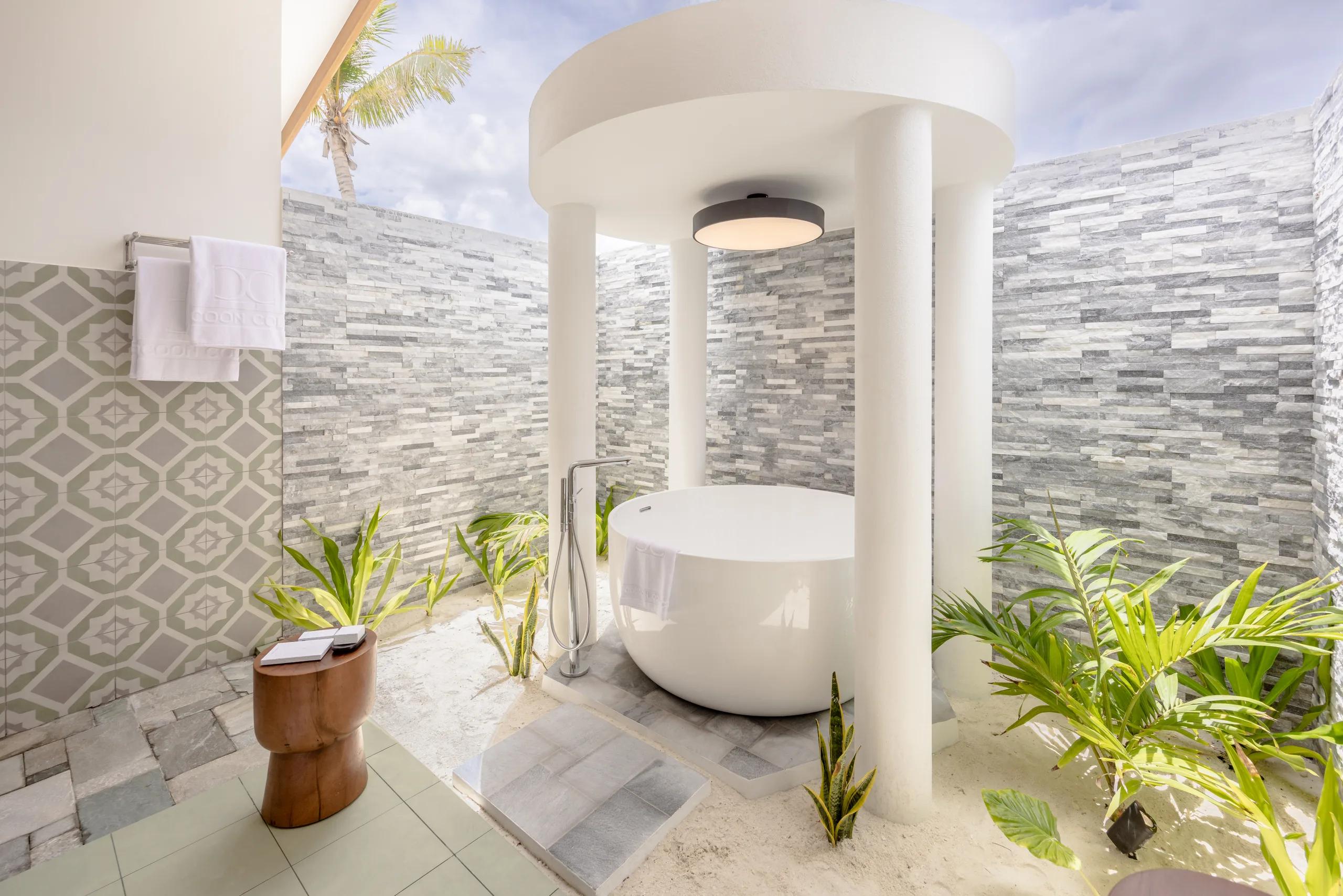 An open-air Maldivian-style bathroom with a freestanding round tub, set among stone walls and palm trees