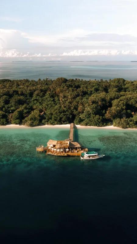 An aerial view of Out of the Sea's rustic shoreline pavilion on stilts, a boat moored alongside and jungle behind