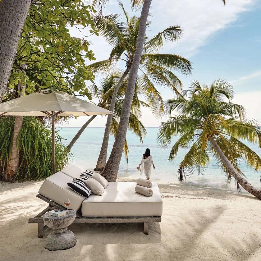Daybed with cushions and towels set up under palm trees on a white-sand beach, a woman walking toward the water