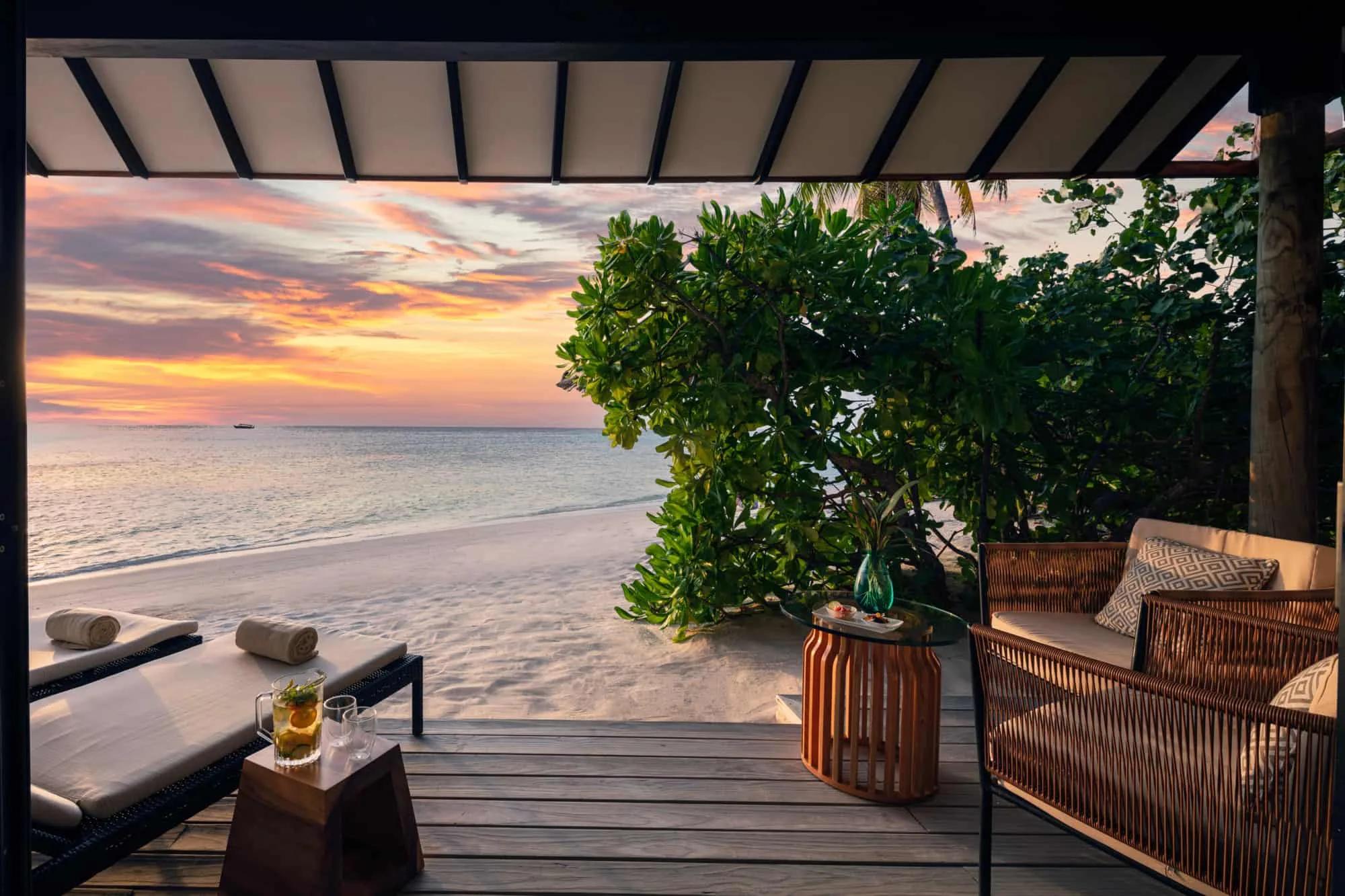 A private terrace at sunset, loungers and a rattan sofa set out on decking beside the beach as the sky turns orange and pink over the sea