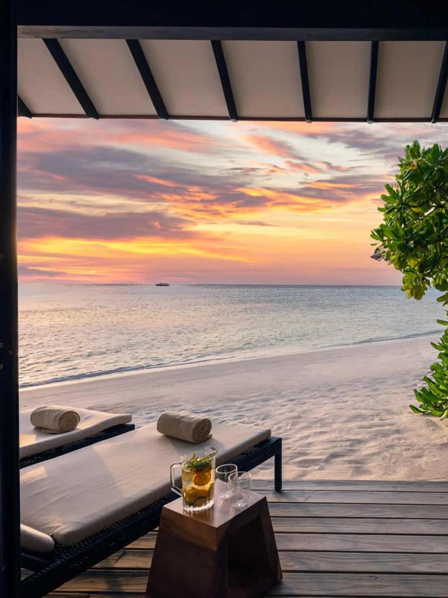 A private terrace at sunset, loungers and a rattan sofa set out on decking beside the beach as the sky turns orange and pink over the sea