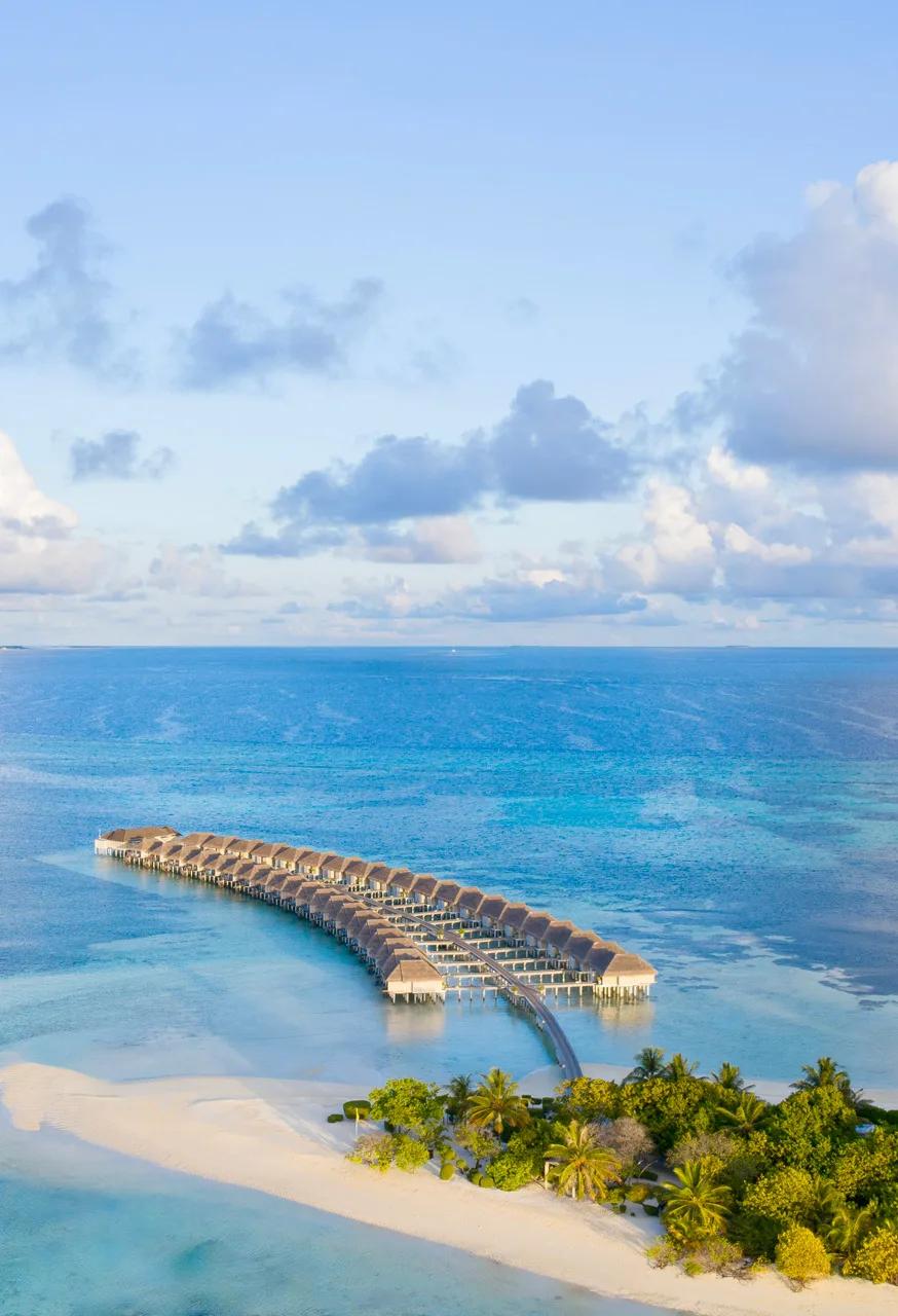 LUX* South Ari Atoll's overwater villa jetty reaching out to the reef edge at golden hour, with the sandy point of the island in the foreground