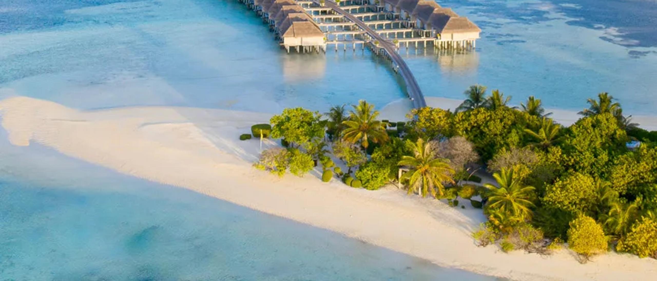 LUX* South Ari Atoll's overwater villa jetty reaching out to the reef edge at golden hour, with the sandy point of the island in the foreground