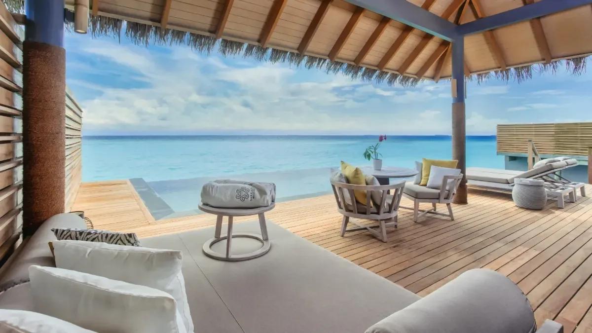 The furnished deck of an Over Water Pool Villa, lounge and dining seating beneath a thatched roof beside the private pool and open lagoon