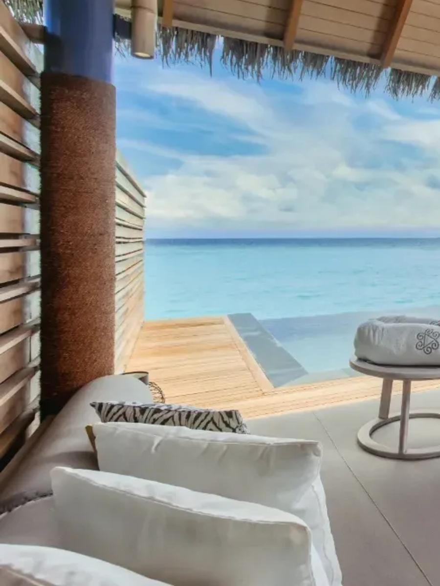 The furnished deck of an Over Water Pool Villa, lounge and dining seating beneath a thatched roof beside the private pool and open lagoon