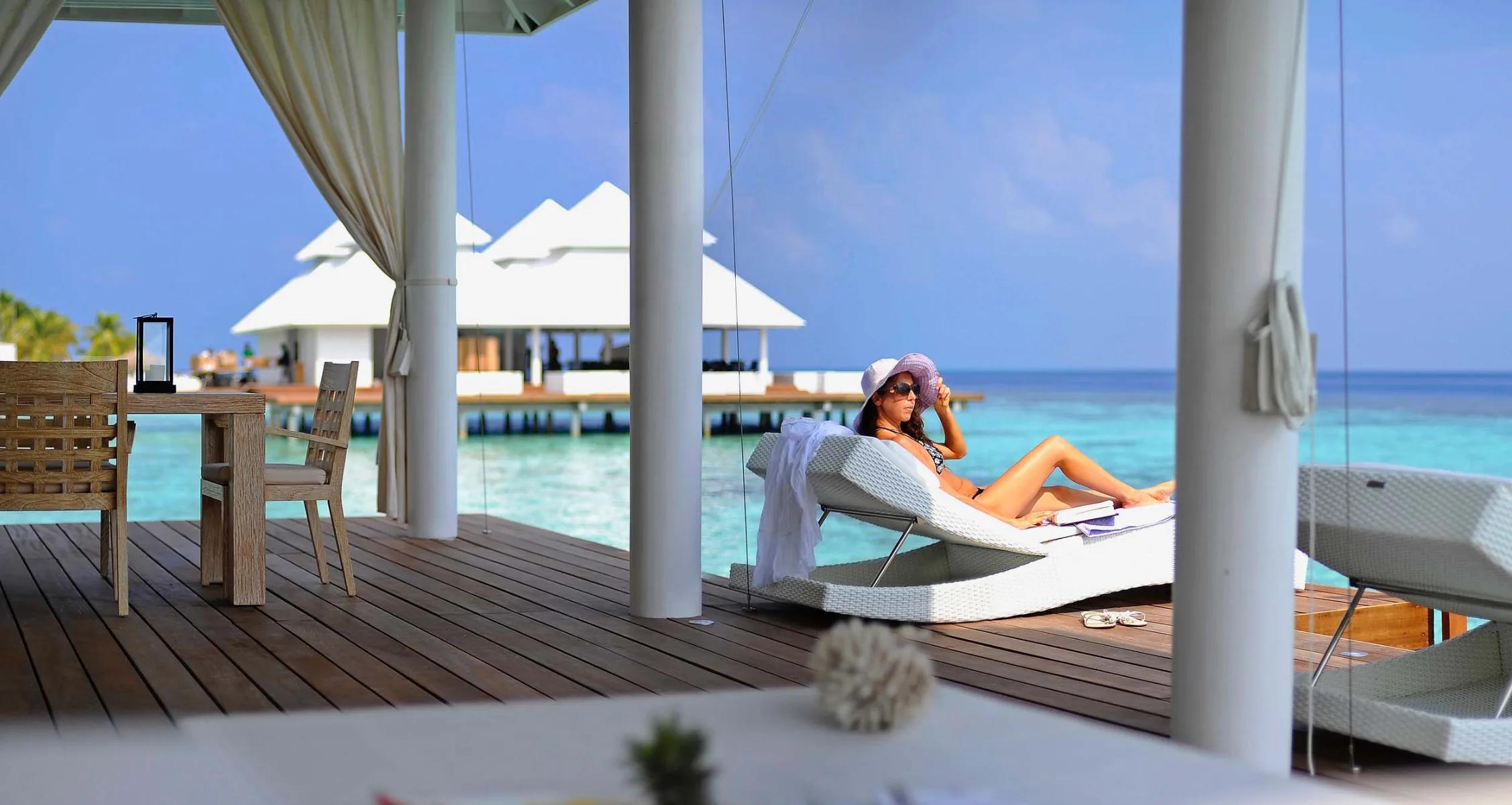 A guest lounging on the deck of a One Bedroom Water Villa at Diamonds Athuruga, water villas visible beyond
