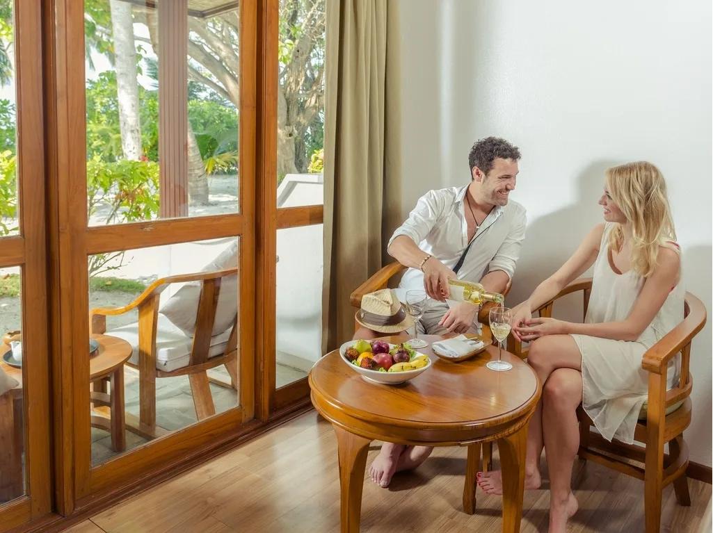 A couple sharing a fruit plate and wine at a table on the veranda of a Standard Beachfront Room, garden visible through the glass doors