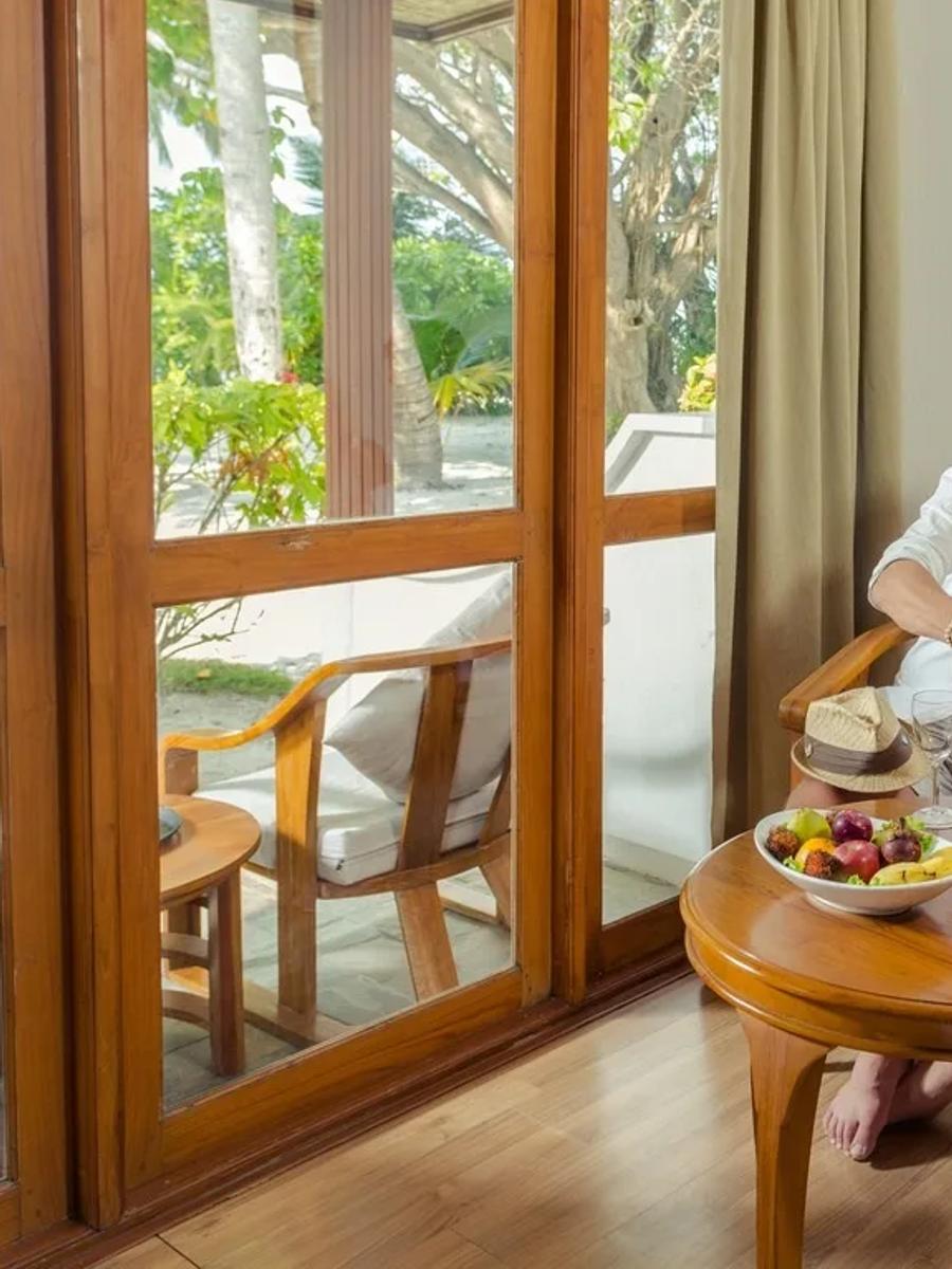 A couple sharing a fruit plate and wine at a table on the veranda of a Standard Beachfront Room, garden visible through the glass doors