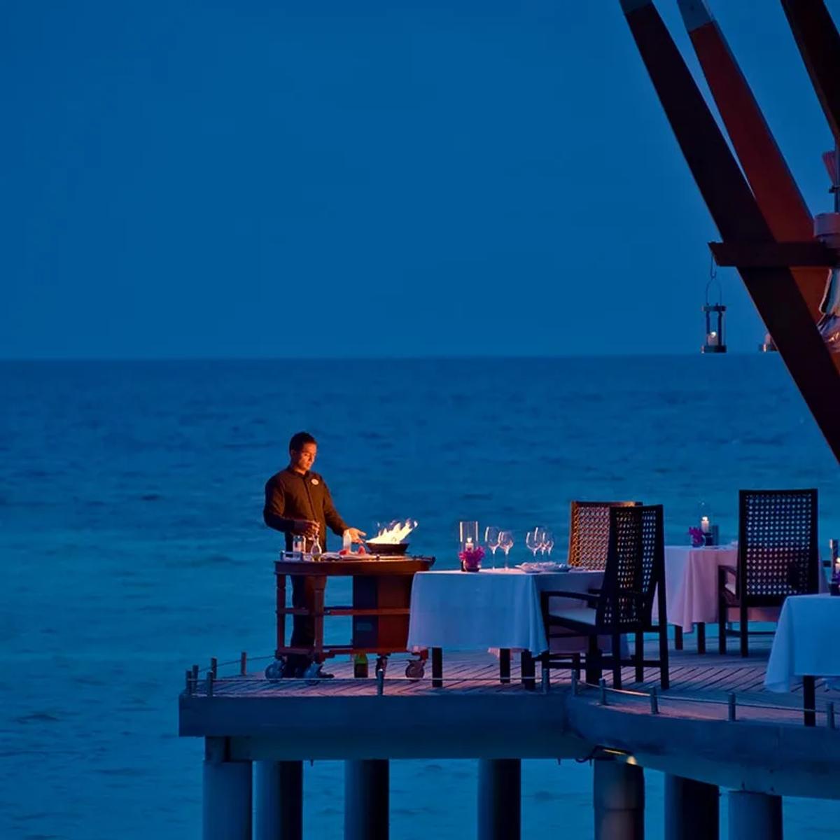 A chef flambéing a dish tableside at The Lighthouse Restaurant, the darkened lagoon visible beyond the overwater deck
