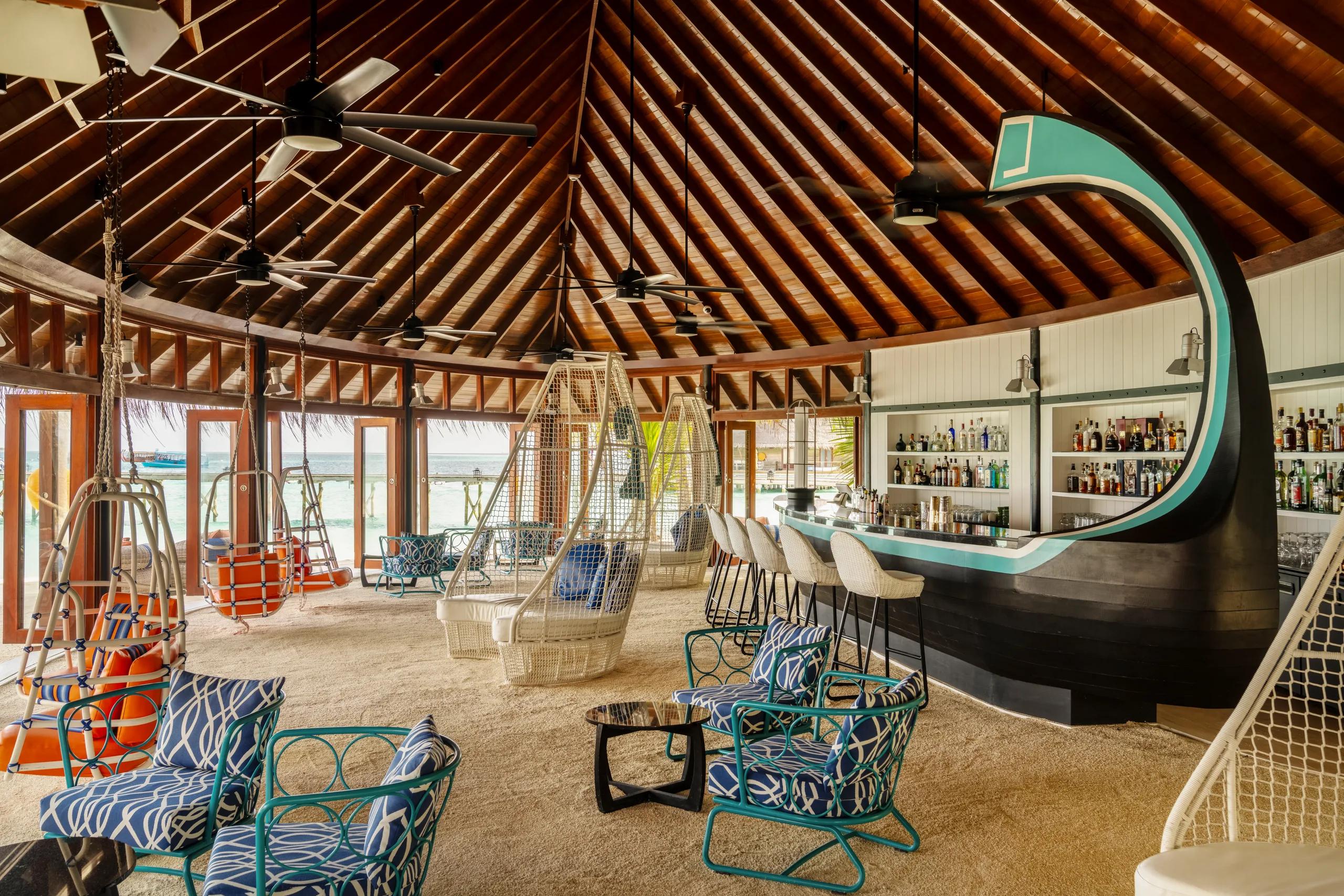 Jahaz Bar & Lounge, its dark boat-shaped counter stocked with spirits, white hanging basket chairs and blue-cushioned seating set on a sand floor