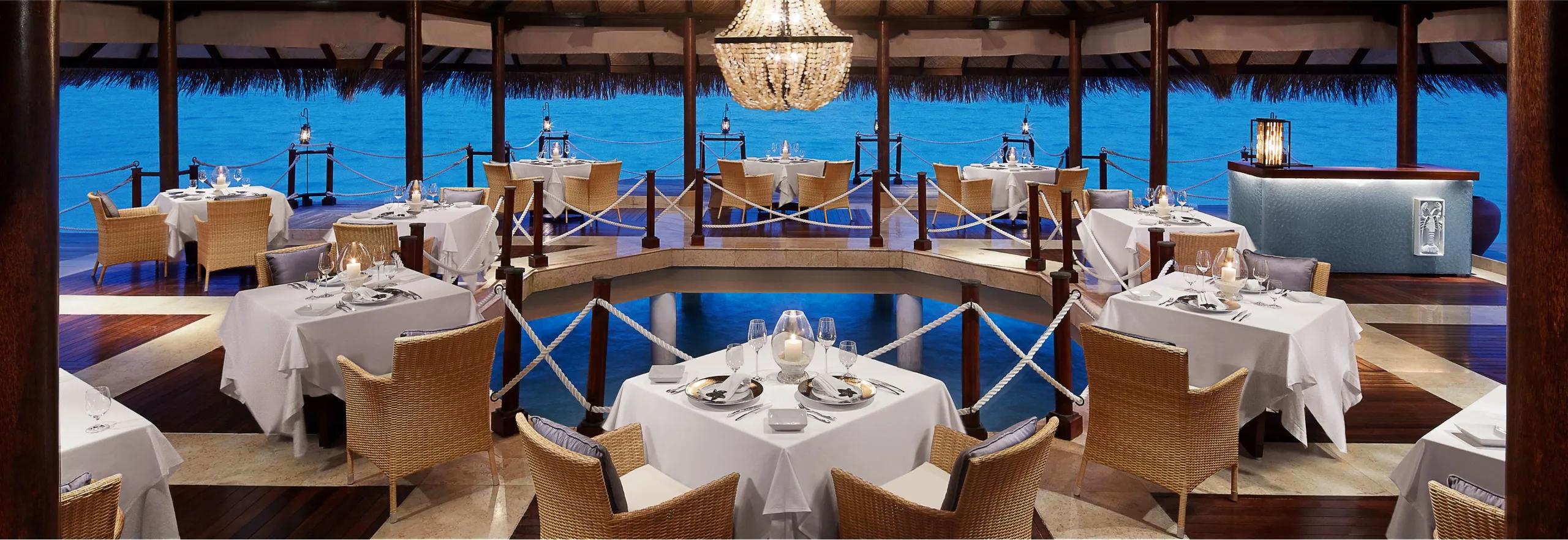 Mediterraneo by Jeffrey Vella's overwater dining room at dusk, tables set beneath a chandelier with the lagoon visible on all sides