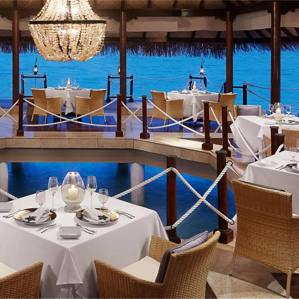 Mediterraneo by Jeffrey Vella's overwater dining room at dusk, tables set beneath a chandelier with the lagoon visible on all sides