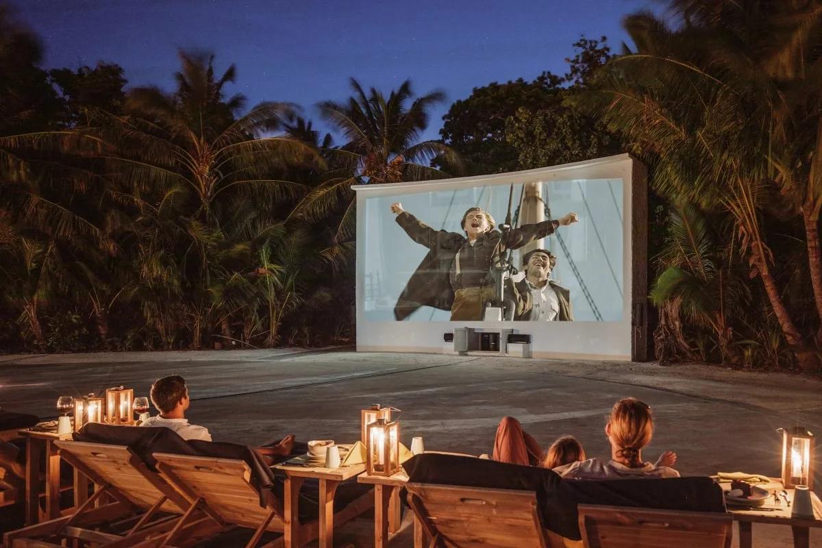 Guests recline on loungers with lantern-lit side tables as a classic film plays on the outdoor screen at Cinema Paradiso, palm trees framing the clearing
