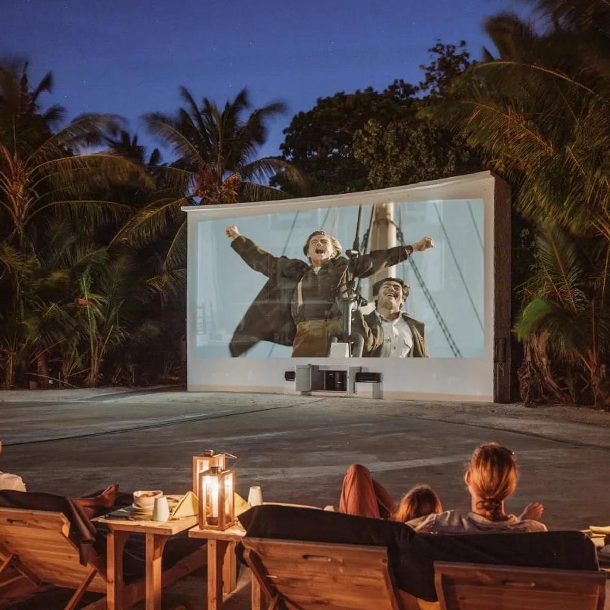 Guests recline on loungers with lantern-lit side tables as a classic film plays on the outdoor screen at Cinema Paradiso, palm trees framing the clearing