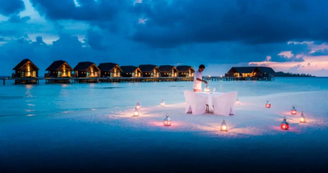 A private candlelit dinner table set on the beach at dusk, with the lit overwater villa row behind it