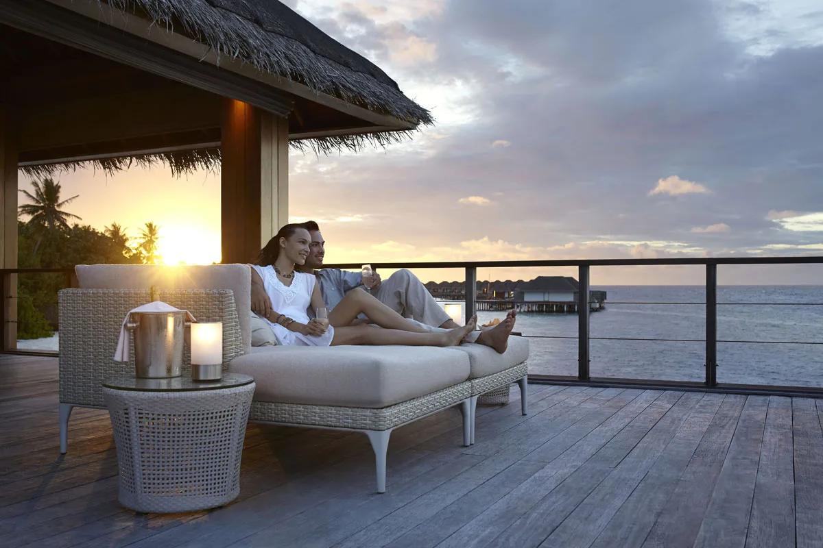 A couple relaxing on an overwater deck at sunset near Sala, Dusit Thani Maldives' plant-based restaurant, overwater villas visible in the distance