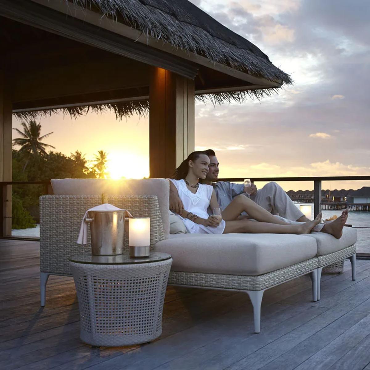 A couple relaxing on an overwater deck at sunset near Sala, Dusit Thani Maldives' plant-based restaurant, overwater villas visible in the distance