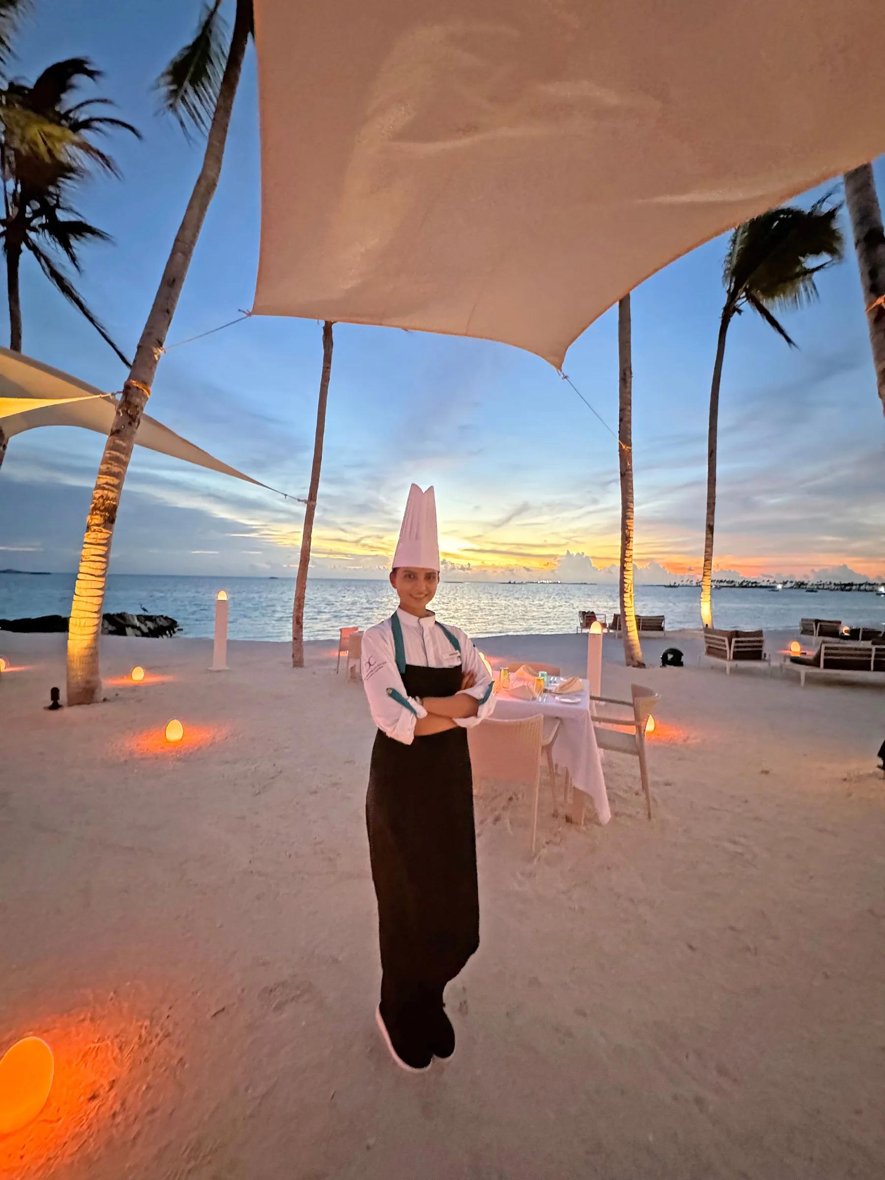 A chef stands beside a private candlelit dinner table set up on the beach at dusk, a canvas canopy and palm trees framing the scene