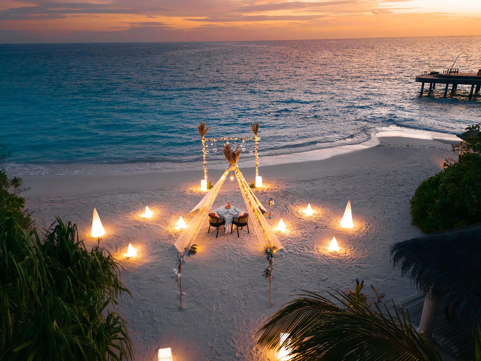 A private beach dinner table for two beneath a lit teepee-style canopy at sunset, cone lanterns arranged across the sand