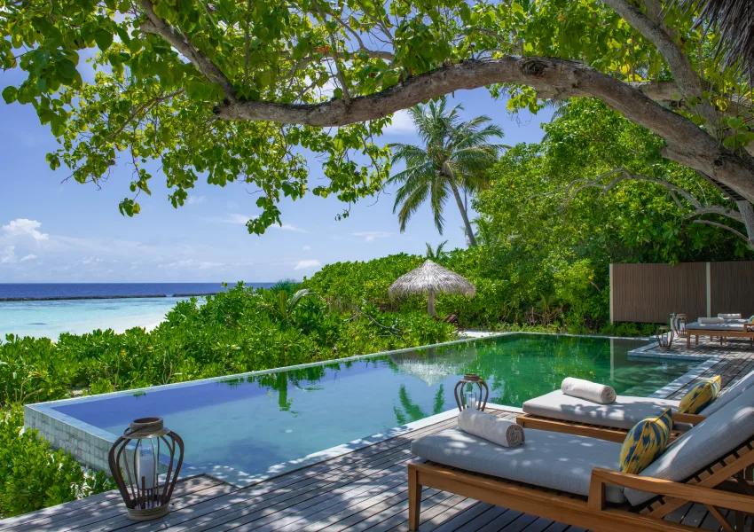 The private pool deck of the Two-bedroom Baros Retreat, shaded by a large tree with the lagoon and reef visible beyond the garden