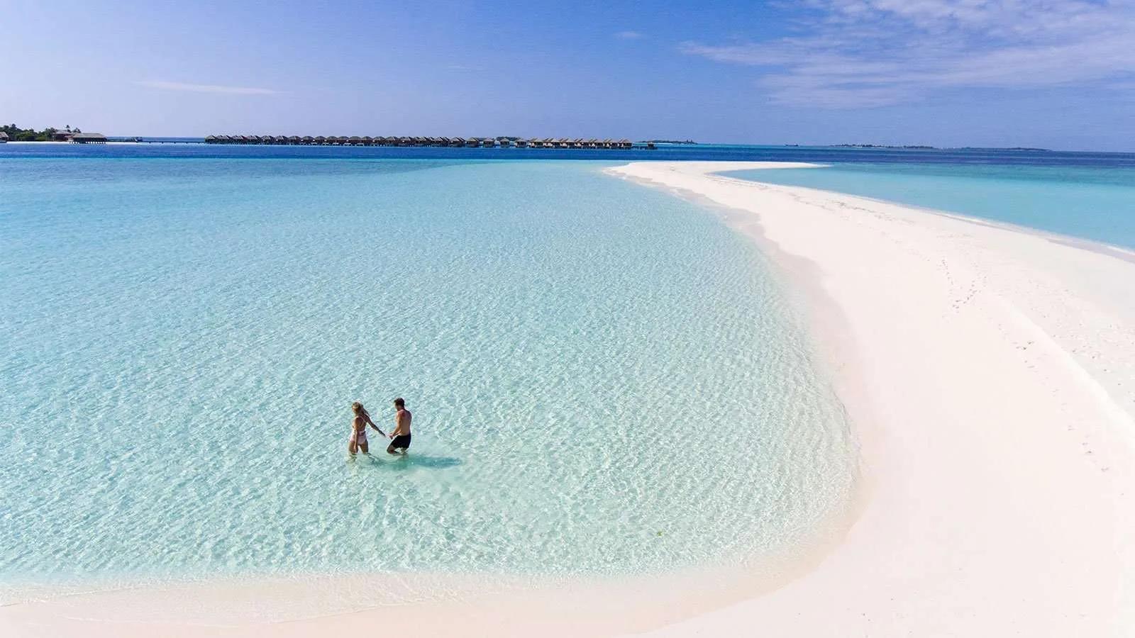 A couple wading hand in hand across a sandbank, a line of overwater villas visible on the horizon