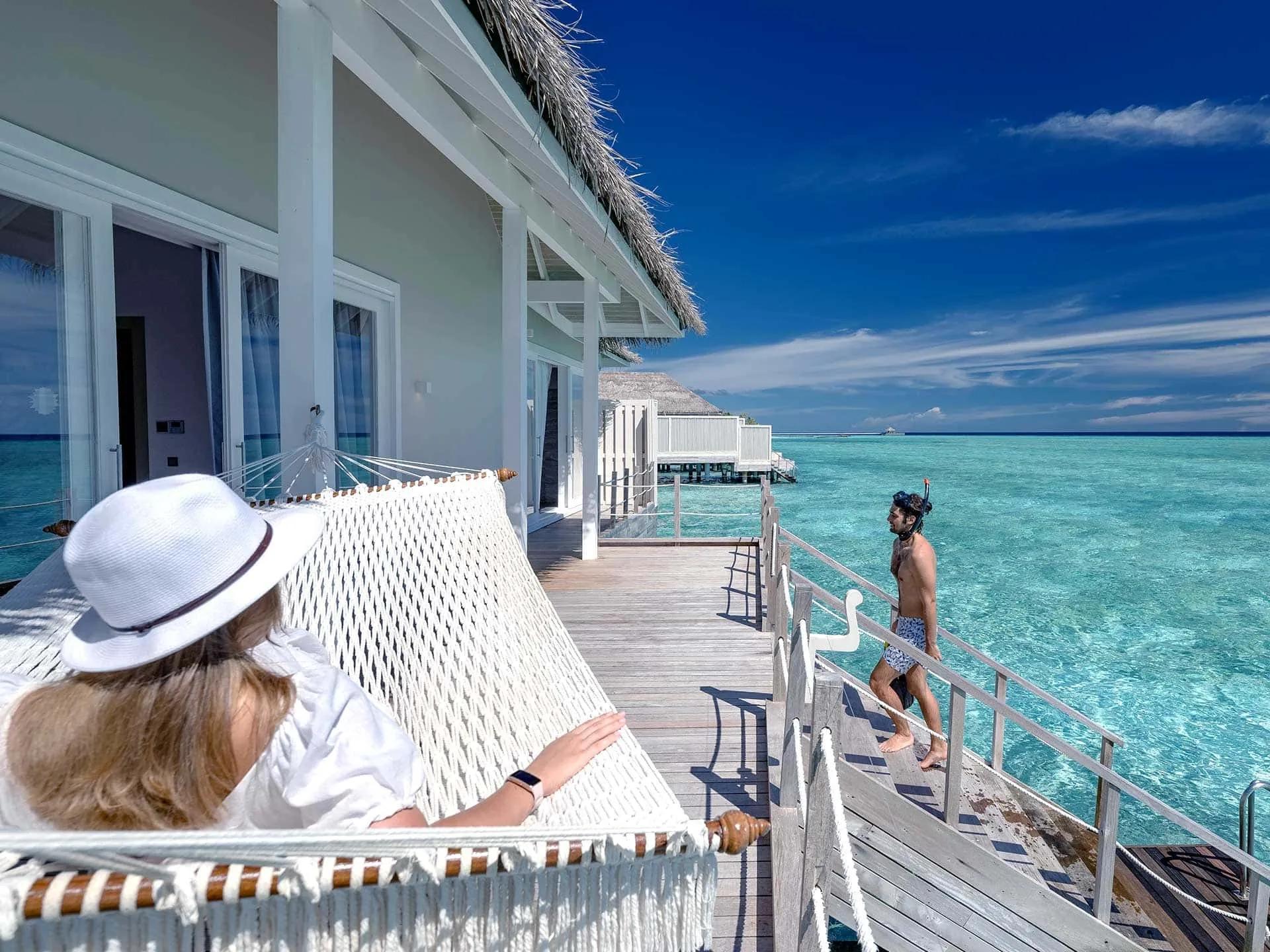 A guest resting in a hammock on the Grand Baglioni Maldives Villa's overwater deck as another walks out in snorkel gear