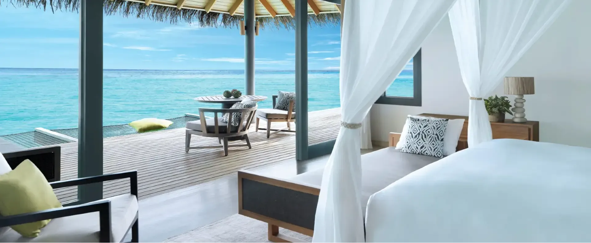 Interior of an Over Water Villa's bedroom and deck, glass doors open to a lounge area and the open lagoon beyond