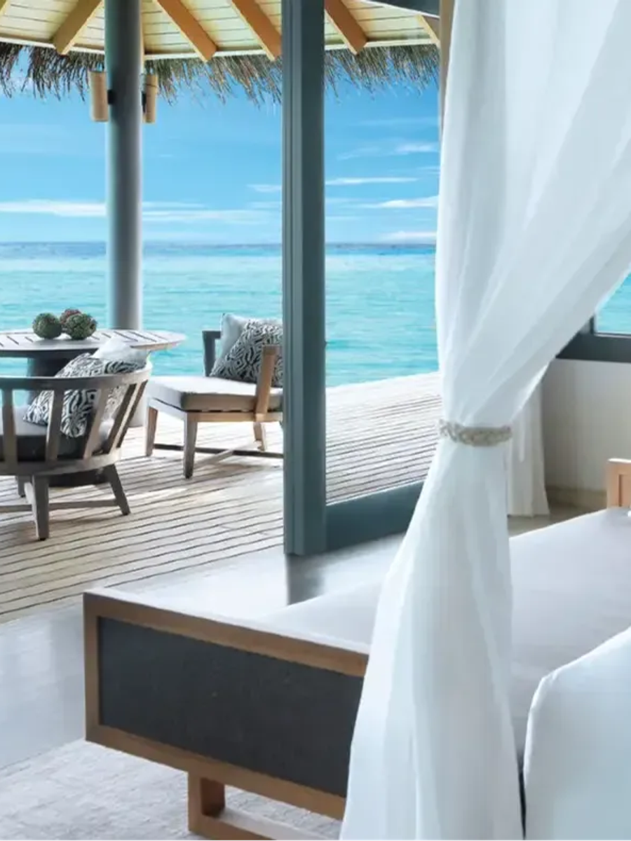 Interior of an Over Water Villa's bedroom and deck, glass doors open to a lounge area and the open lagoon beyond