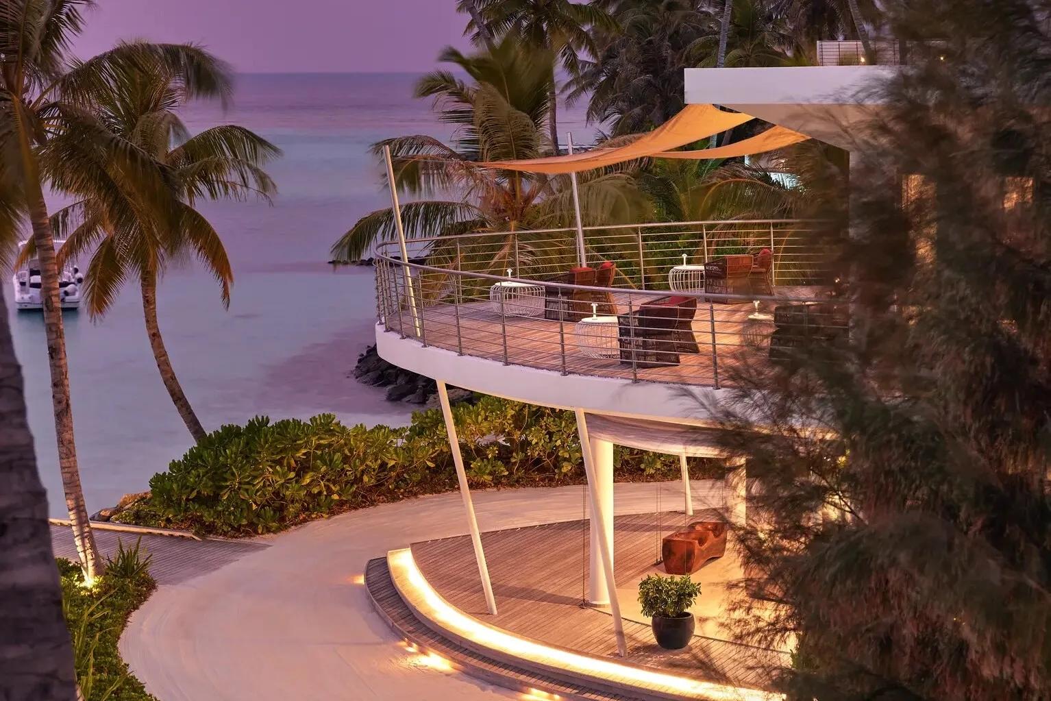 An elevated curved terrace with lounge seating overlooking the ocean at dusk, a boat moored offshore