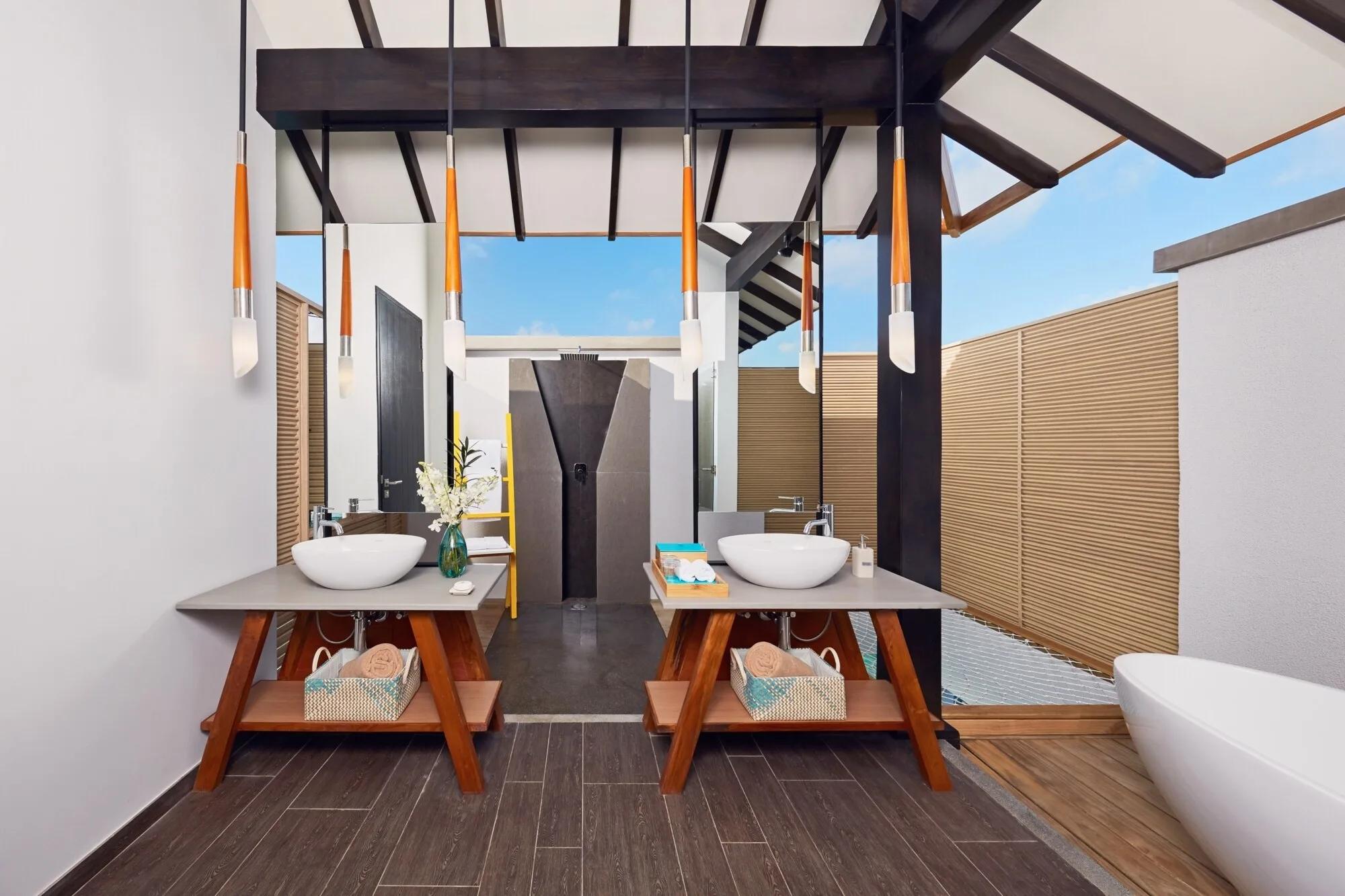 An open-air double-vanity bathroom in the Over Water Pool Suite, pendant lights hanging from a timber-beamed roof and a freestanding tub beside a slatted privacy screen