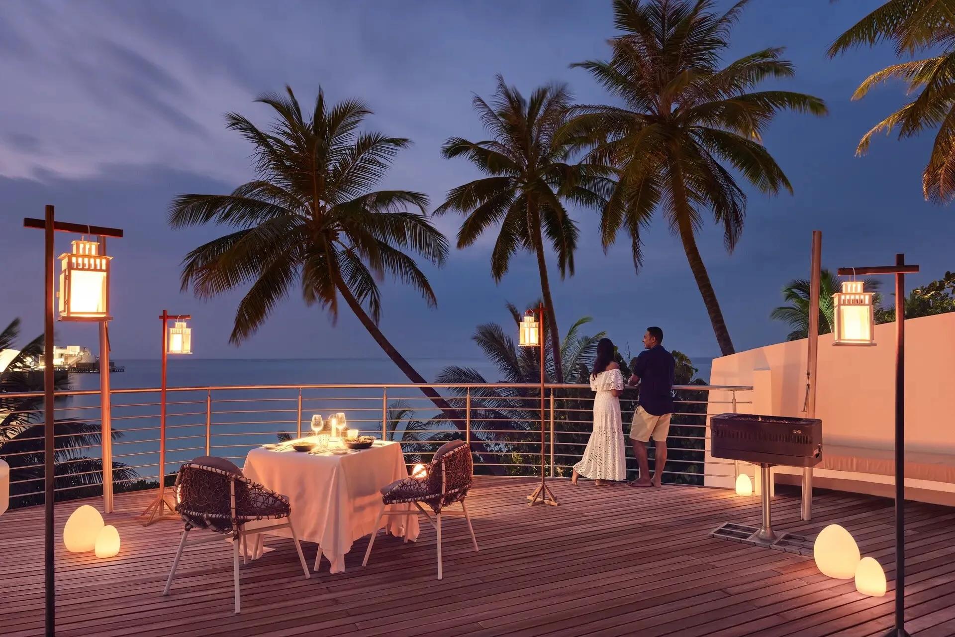 A private dinner set on a villa's roof terrace at dusk, lanterns lit and the overwater villa row visible across the water