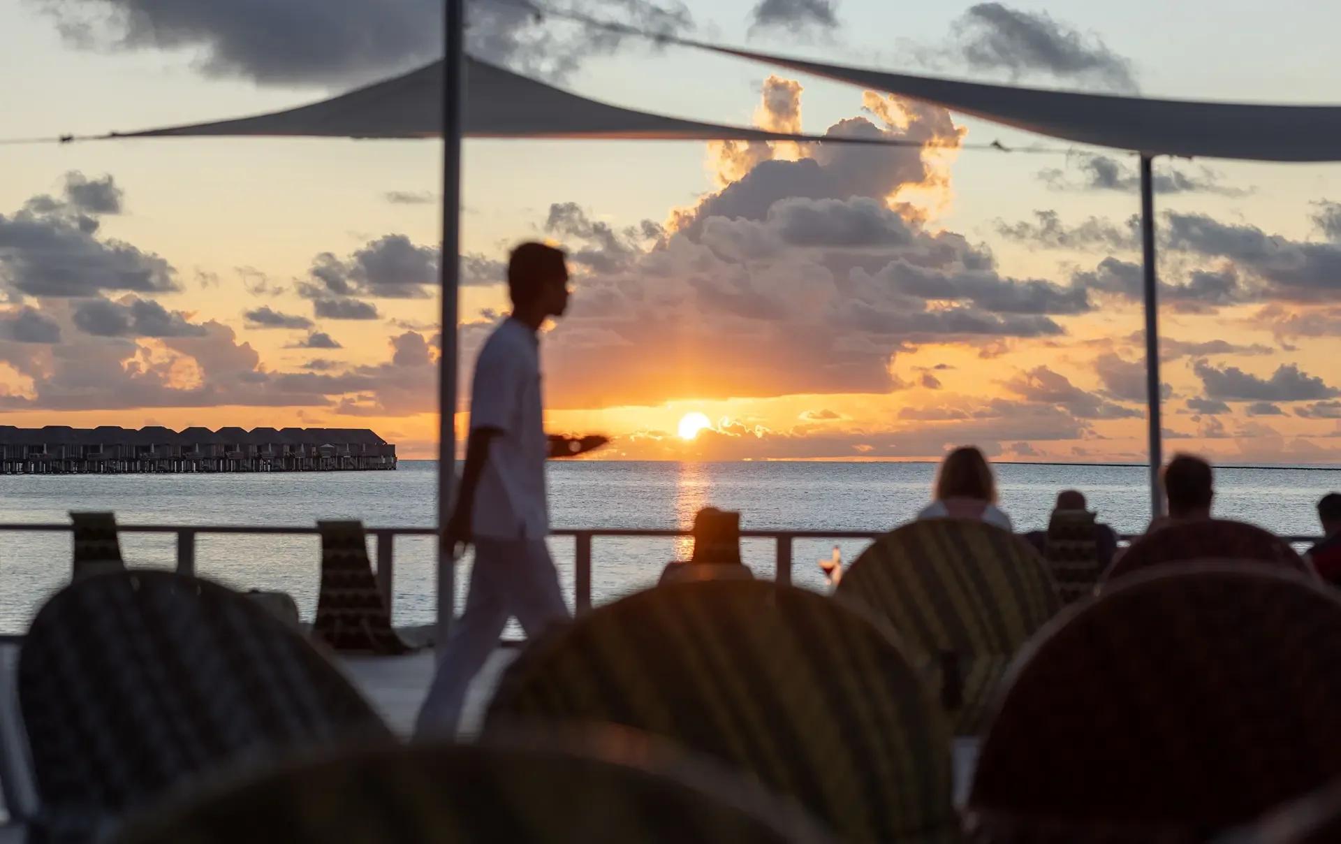 A waiter carrying drinks past silhouetted guests at Chill Bar, with a row of overwater villas visible across the lagoon at sunset