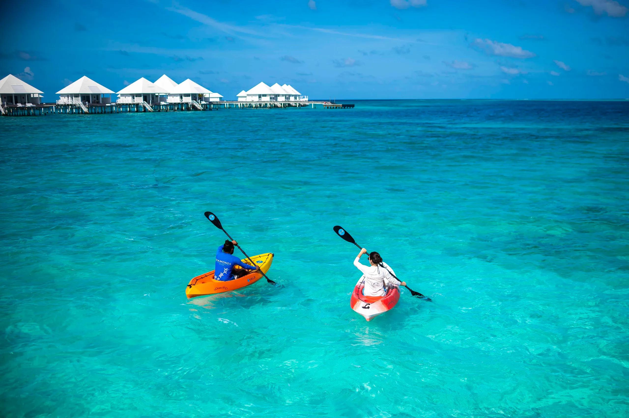 Two guests kayaking across the lagoon toward Diamonds Thudufushi's white-roofed overwater villas